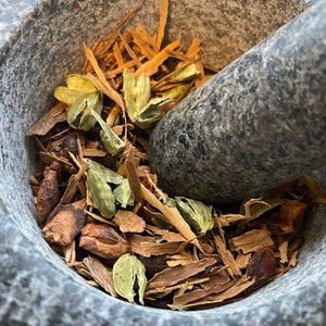 May include: A close-up of a gray stone mortar and pestle with a mixture of brown cinnamon sticks, green cardamom pods, and other spices inside.