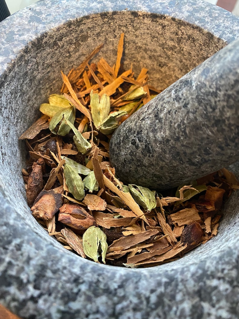 May include: A close-up of a gray stone mortar and pestle with a mixture of brown cinnamon sticks, green cardamom pods, and other spices inside.