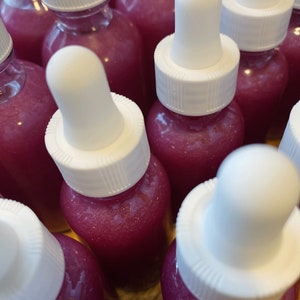 K&ouml;nnte beinhalten: Eine Gruppe kleiner Glasflaschen mit wei&szlig;en Tropfern, gef&uuml;llt mit einer schimmernden rosa Fl&uuml;ssigkeit. Die Flaschen sind in einer Nahaufnahme angeordnet.
