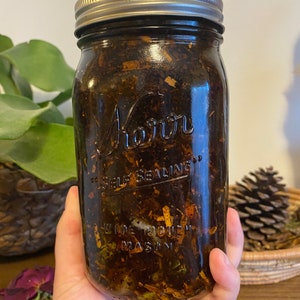 May include: A clear glass jar with a silver lid filled with a dark brown liquid and various herbs and plant materials. The jar is labeled "Kerr" and "Self Sealing".
