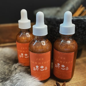 May include: Three amber glass bottles with white dropper tops and labels that say "Sun Summoner Spiced Coffee + Cacao Face & Body Shimmer Serum 1 fl oz".
