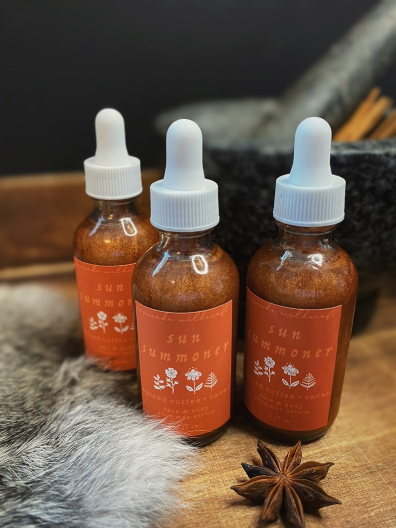 May include: Three amber glass bottles with white dropper tops and labels that say "Sun Summoner Spiced Coffee + Cacao Face & Body Shimmer Serum 1 fl oz".