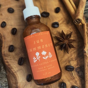 May include: A glass bottle with a gold shimmery liquid inside. The bottle has a white dropper and a label that says "anade wilderaft sun summoner spiced coffee + cacao face & body shimmer serum 2 fl oz".