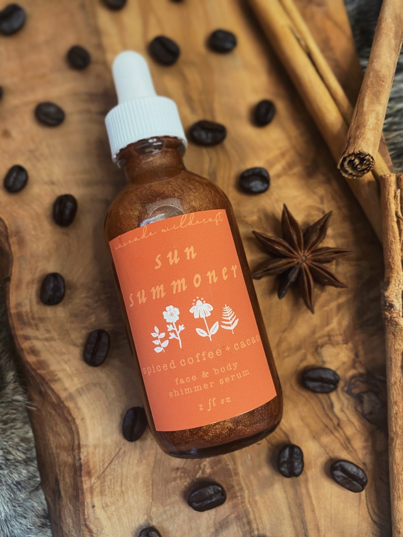 May include: A glass bottle with a gold shimmery liquid inside. The bottle has a white dropper and a label that says "anade wilderaft sun summoner spiced coffee + cacao face & body shimmer serum 2 fl oz".