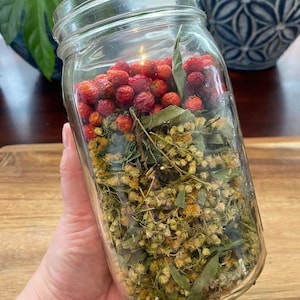 May include: A glass jar filled with dried herbs and red berries. The jar is clear and shows the contents inside. The jar is being held by a hand.