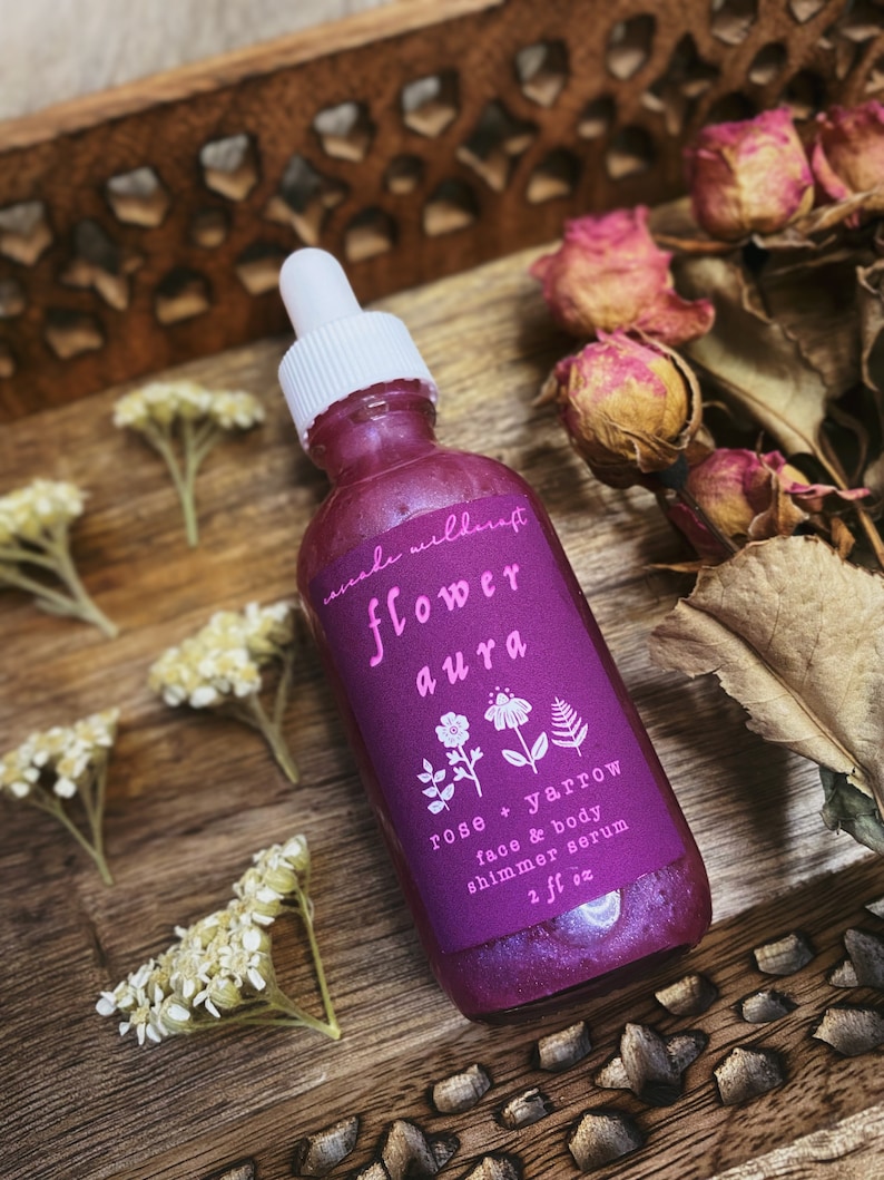 K&ouml;nnte beinhalten: Eine violette Glasflasche mit einem wei&szlig;en Tropfer und einem rosa Etikett mit der Aufschrift "made wildcraft flower aura rose + yarrow face & body shimmer serum 2 fl oz".