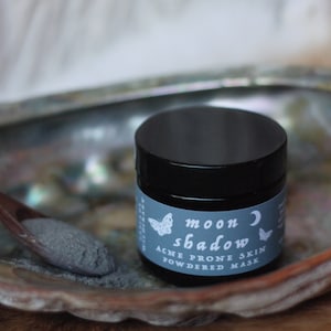 May include: A black jar with a blue label that says "moon shadow acne prone skin powdered mask" with a white crescent moon and butterfly graphic.