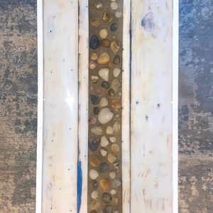 Puede incluir: Una bandeja de madera blanca con una incrustación de resina transparente que presenta una línea vertical de conchas y pequeñas piedras. La bandeja tiene un marco pintado de blanco.