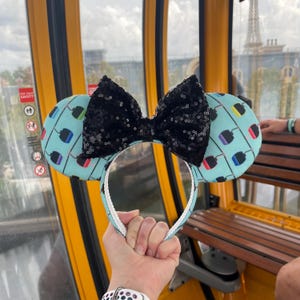 May include: A pair of blue and white mouse ears with a black sequin bow. The ears feature a pattern of colorful light bulbs.