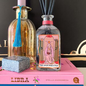 Könnte beinhalten: Eine rosa Kerze in einem goldenen Kerzenhalter mit einem blauen Quasten, eine klare Glasflasche mit einem roten Deckel und einem Tarotkartenaufkleber der Hohenpriesterin, ein grauer Stein und eine rosa und blaue Schachtel mit den Worten "LIBRA" und "LEO" darauf.