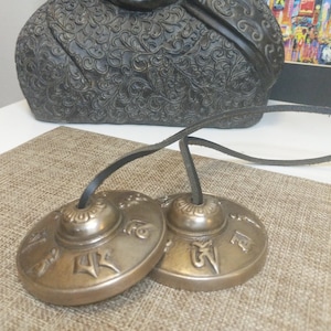 May include: Two small, round, brass Tibetan singing bowls with intricate designs and Sanskrit lettering. The bowls are connected by a black leather cord.