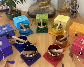 Puede incluir: Siete cuencos cantores con mazos de madera, cada uno de un color diferente del arcoíris, dispuestos en círculo sobre una mesa de madera. Los cuencos están acompañados de sus cajas de chakras correspondientes, cada una de un color y símbolo diferente. Una estatua dorada de Buda se encuentra en el fondo, junto con un póster de los siete chakras.