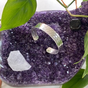 May include: A silver hammered cuff bracelet resting on a bed of purple amethyst crystals.