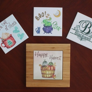 May include: Four decorative ceramic tiles with various designs. One tile features a mug with the text "Have a cup of cheer". Another says "Boo! 2 Stew" with a cauldron. A third tile has the name "Blackwell". The last tile says "Happy Harvest" with pumpkins.