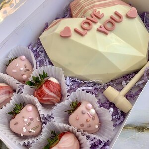 Puede incluir: Una caja de chocolate blanco en forma de coraz&oacute;n con las palabras "Love You" en letras rosas. La caja contiene un mazo de madera y seis fresas cubiertas de chocolate con chispas rosas y blancas.