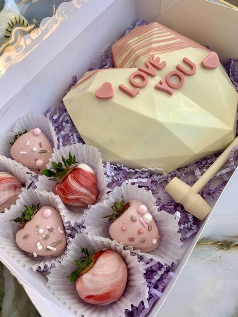 Puede incluir: Una caja de chocolate blanco en forma de coraz&oacute;n con las palabras "Love You" en letras rosas. La caja contiene un mazo de madera y seis fresas cubiertas de chocolate con chispas rosas y blancas.