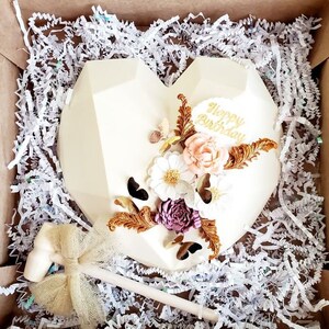 Puede incluir: Pastel en forma de coraz&oacute;n de chocolate blanco decorado con letras doradas que dicen "Happy Birthday" y detalles florales blancos, rosas y dorados. El pastel est&aacute; en una caja con papel blanco triturado.