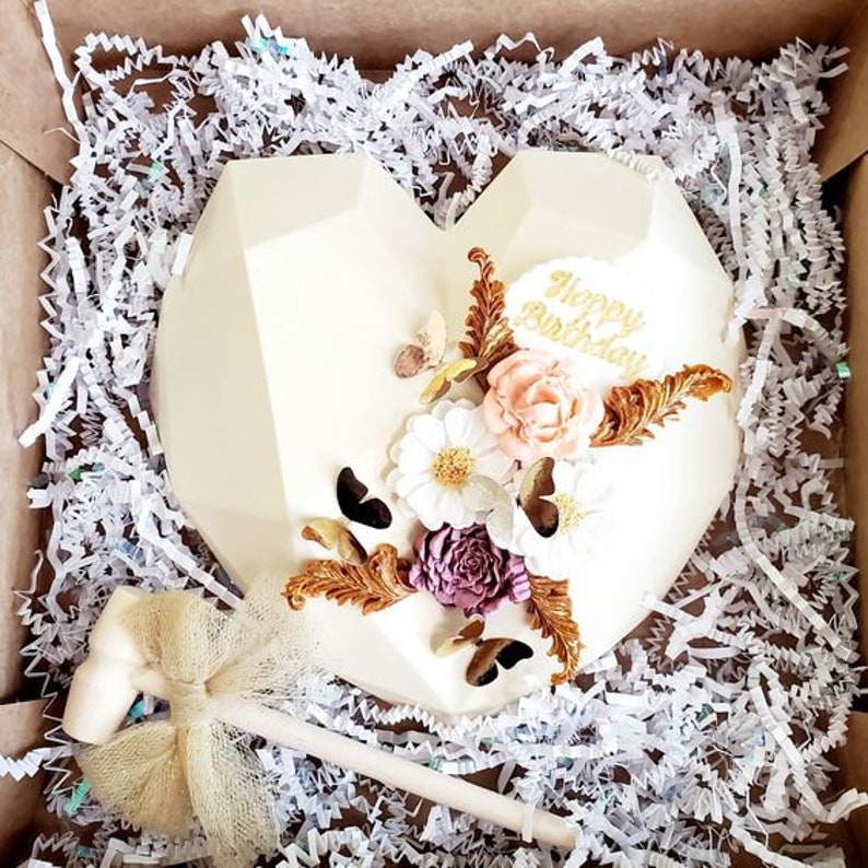 Puede incluir: Pastel en forma de coraz&oacute;n de chocolate blanco decorado con letras doradas que dicen "Happy Birthday" y detalles florales blancos, rosas y dorados. El pastel est&aacute; en una caja con papel blanco triturado.