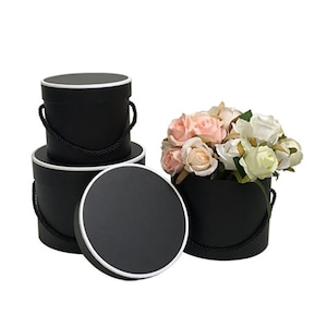 May include: Three black round hat boxes with white trim and black rope handles. The top box is empty, the middle box is empty, and the bottom box contains a bouquet of pink and white roses.