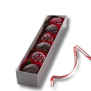 May include: A white box filled with chocolate-covered strawberries, each topped with pink and white sprinkles. The box is open and a red ribbon is lying next to it.