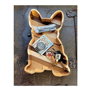 May include: A wooden tray shaped like a French bulldog, with a multi-colored wood grain pattern. The tray is filled with various items, including a multi-tool, a dollar bill, a coin, and a set of keys.