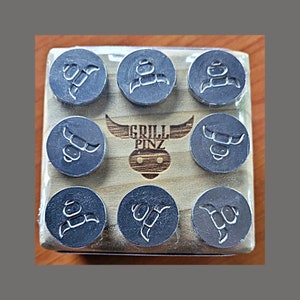 May include: A set of eight black metal grill pinz with different designs. The pinz are arranged in a grid on a wooden board with the text "Grill Pinz" engraved in the center.