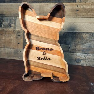 Pu&ograve; includere: Un vassoio di servizio in legno a forma di cane con una finitura in legno naturale. Il vassoio &egrave; inciso con i nomi "Bruno & Bella".