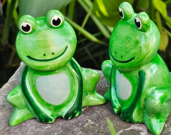 May include: Two green ceramic frog figurines with white eyes and white bellies. The frogs are sitting on a grey stone surface.
