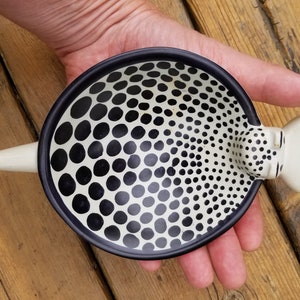 Puede incluir: Un plato de cer&aacute;mica blanco con un patr&oacute;n de lunares negros y blancos. El plato tiene un peque&ntilde;o pico redondeado blanco en un lado.