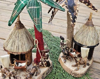 May include: Two miniature thatched hut villages with palm trees, one with a green and white plaid palm tree and the other with a brown palm tree. The huts are made of straw and have small figures of people around them.