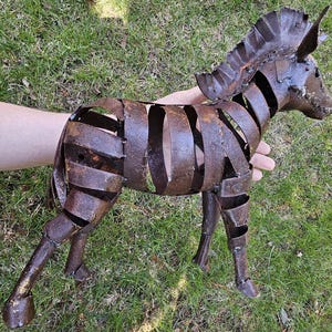 Puede incluir: Escultura de cebra de metal oxidado con dise&ntilde;o de rayas. La escultura est&aacute; hecha de tiras de metal soldadas, creando una forma abierta tridimensional. La cebra est&aacute; de pie sobre cuatro patas, con cabeza y melena detalladas. La escultura es de color marr&oacute;n desgastado.