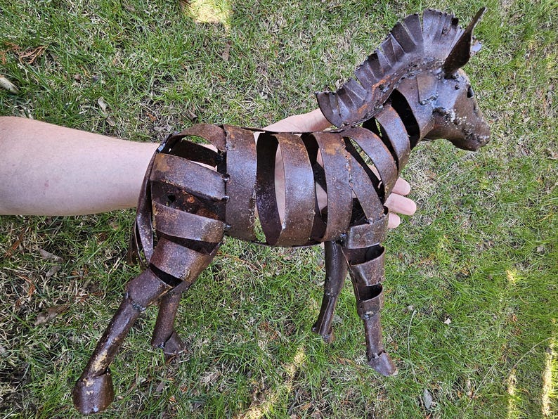 Puede incluir: Escultura de cebra de metal oxidado con dise&ntilde;o de rayas. La escultura est&aacute; hecha de tiras de metal soldadas, creando una forma abierta tridimensional. La cebra est&aacute; de pie sobre cuatro patas, con cabeza y melena detalladas. La escultura es de color marr&oacute;n desgastado.