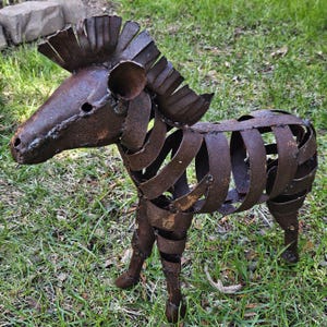 Puede incluir: Escultura de metal oxidado que representa un cebra. El cuerpo del animal est&aacute; formado por tiras de metal curvadas, con una cabeza y una melena detalladas. La escultura se encuentra sobre una superficie cubierta de hierba.
