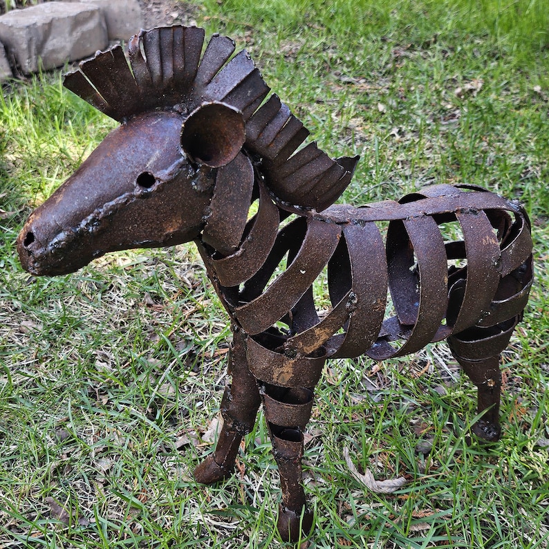Puede incluir: Escultura de metal oxidado que representa un cebra. El cuerpo del animal est&aacute; formado por tiras de metal curvadas, con una cabeza y una melena detalladas. La escultura se encuentra sobre una superficie cubierta de hierba.