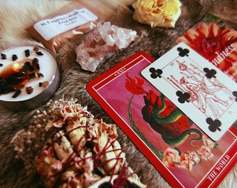 Può includere: Una collezione che include una carta dei tarocchi rossa con l'etichetta The World, una carta da gioco sei di fiori, una candela accesa con inclusioni scure, un pacchetto con l'etichetta St. Catherine's Monastery Brick Dust, quarzo e fiori secchi.