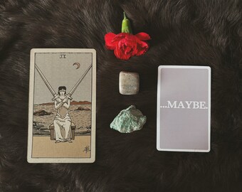 Könnte beinhalten: Eine Zwei-der-Schwerter-Tarotkarte mit einer verbundenen Figur, die zwei gekreuzte Schwerter hält, eine rote Nelke, einen glatten beigen Stein, ein raues grünes Mineral und eine rechteckige Karte mit dem Text ...MAYBE. in Weiß.