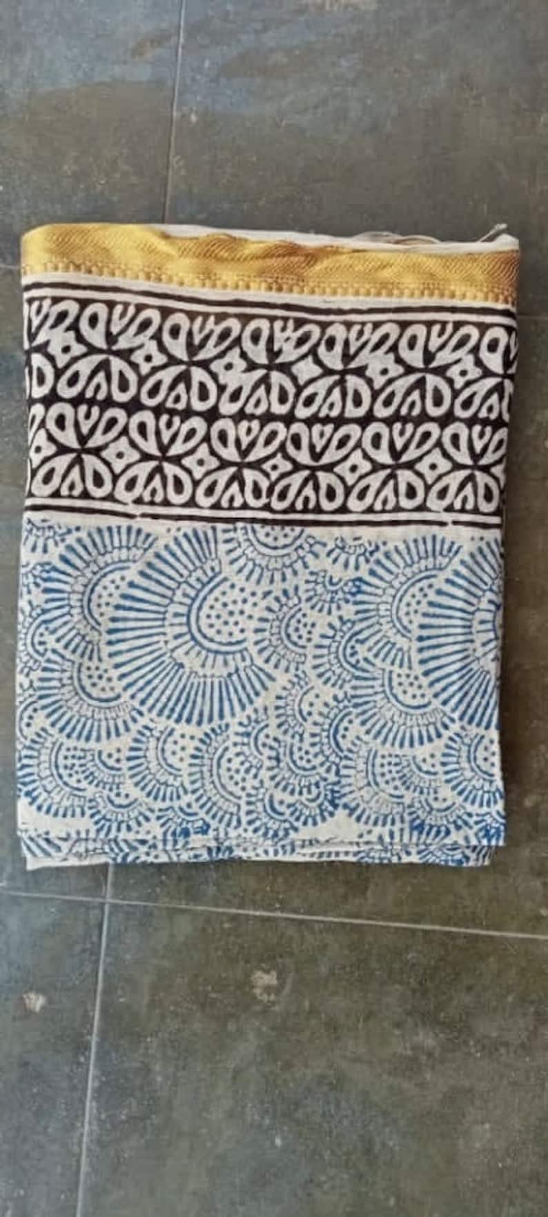 May include: A white cotton scarf with a blue and white paisley pattern. The scarf has a gold trim around the edges.