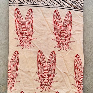 Könnte beinhalten: Ein gefaltetes, cremefarbenes Textil mit einem sich wiederholenden Muster aus roten Zikaden. Der obere Rand hat eine schwarze geometrische Bordüre. Die Zikaden sind mit Flügel- und Körpermustern detailliert, was einen auffälligen Kontrast erzeugt.