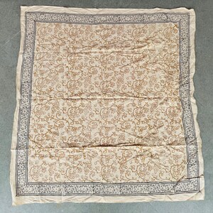 May include: A square, cream-colored textile with a repeating floral pattern in brown. The border features a dark blue and white decorative design. The fabric is slightly wrinkled, suggesting a soft material. This textile could be a tablecloth or a decorative throw.