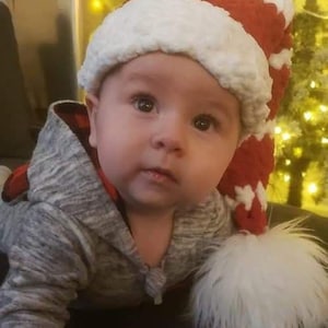 May include: A baby wearing a red and white crocheted Santa hat with a white fur trim, lying on a black surface. The baby is wearing a grey hooded sweater.