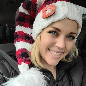 May include: A woman wearing a red, white, and black crocheted hat with a white pom-pom. The hat has a wooden button with a star design.