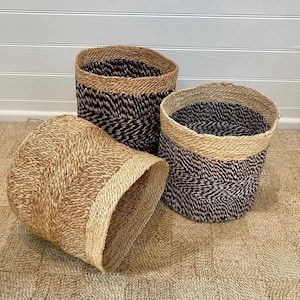 May include: Three woven storage baskets in different sizes. The baskets are in natural tan with black and tan patterned details. The rims of the baskets are tan. The baskets are on a woven mat.