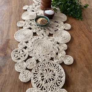 Peut inclure: Chemin de table en fibre naturelle avec un motif circulaire. Le chemin de table est fait de corde étroitement enroulée, créant une surface texturée. Le motif présente des cercles et des motifs floraux de différentes tailles. Il est posé sur une table en bois, avec des petits bols et un moulin à poivre en bois.