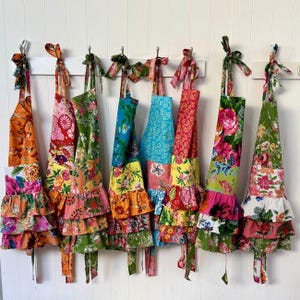 May include: A collection of colorful patchwork aprons hanging on a white wall. Each apron features a unique floral pattern with ruffled hems and matching ties. The aprons are in shades of orange, pink, green, blue, and red.