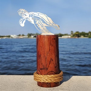 May include: A white metal sea turtle sculpture mounted on a brown wooden cylinder with a rope wrapped around the base. The sculpture is set against a blue water background.