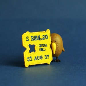 May include: A yellow felt patch with the text "S RM4-20 GUNA 981 31 AUG 57" in blue embroidery. A brown kiwi bird figurine is next to the patch.