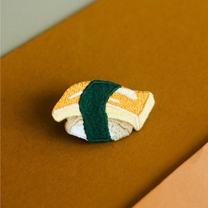 Pode incluir: Um remendo de sushi bordado em amarelo e branco com um invólucro de alga verde. O remendo está sobre uma superfície castanha.