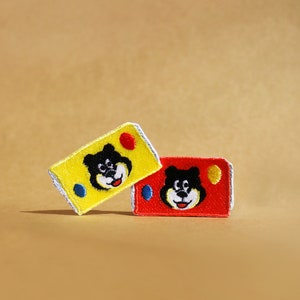 Może przedstawiać: Dwa haftowane naszywki z postaciami kreskówkowych niedźwiedzi z czarnymi oczami i nosami. Naszywka po lewej stronie jest żółta z czerwonym kółkiem i niebieskim kółkiem. Naszywka po prawej stronie jest czerwona z niebieskim kółkiem i białym kółkiem.