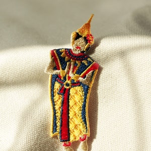 May include: Embroidered patch of a person in a yellow, red, and blue costume. The person is holding a staff in their hands.