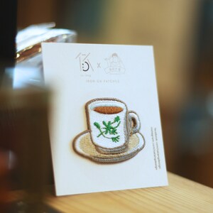 Op de afbeelding: Een witte kaart met een bruin geborduurd strijk-op-patch van een kopje thee met groene bladeren. De kaart heeft de tekst "Iron On Patches" en een logo met de tekst "13 x 鹹魚仔".